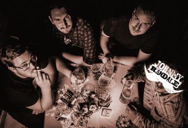 Bandfoto Johnny Randale: Vier Personen sitzen an einem Tisch, der mit Spielkarten, Gläsern und Flaschen bedeckt ist. Das Foto ist von oben aufgenommen und in dunklen, kontrastreichen Farbtönen gehalten. Die Personen schauen unterschiedlich in die Kamera o